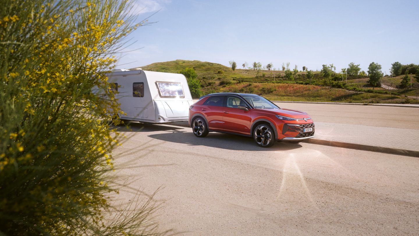 Ein roter T-Roc zieht einen weißen Wohnwagen auf einer asphaltierten Straße. Die Landschaft ist sonnig und zeigt grüne Hügel im Hintergrund. Ein Strauch mit gelben Blüten ist an der linken Seite des Bildes zu sehen.