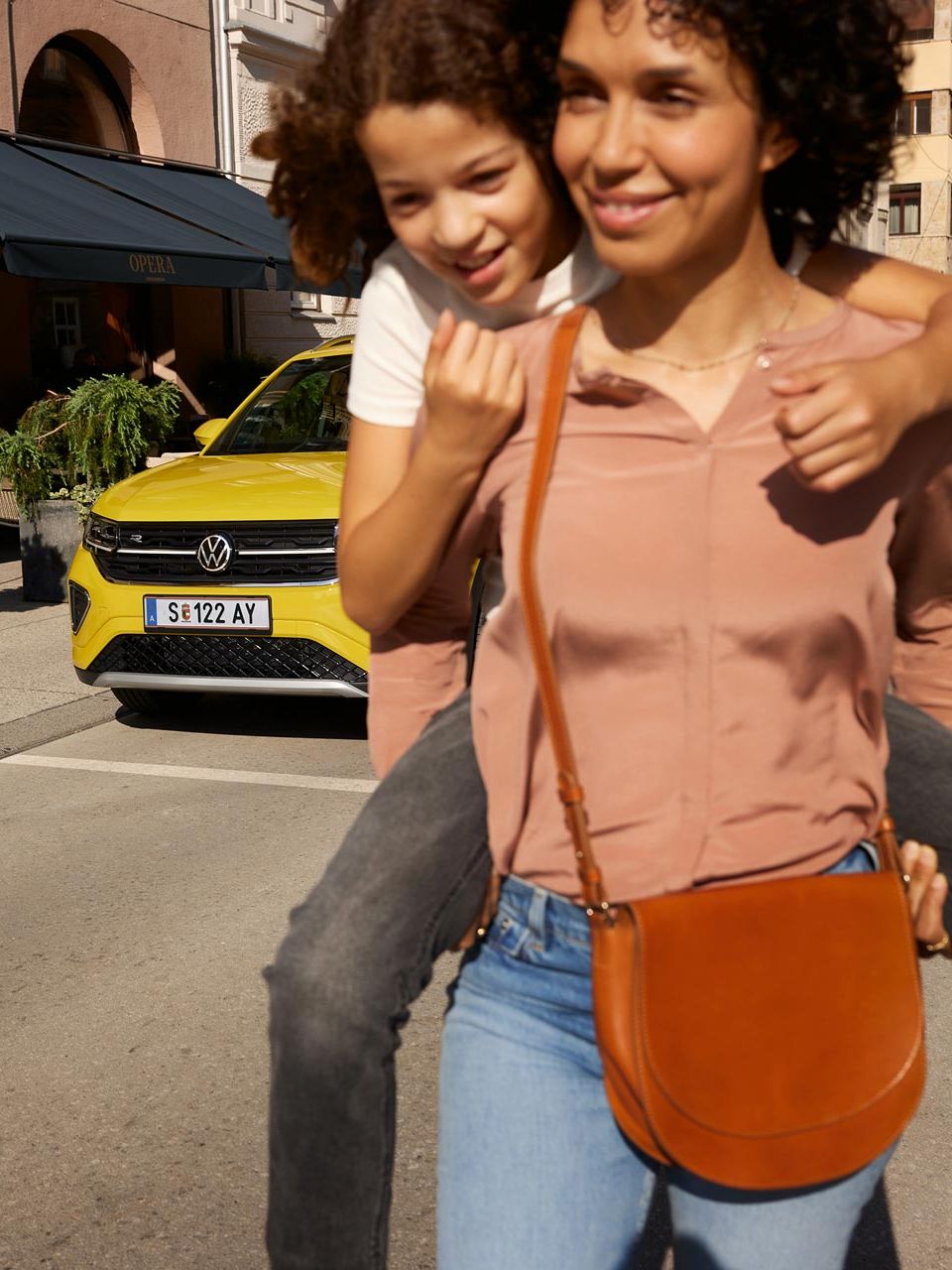 Auf einer Straße in der Innenstadt trägt eine Frau ein Mädchen auf dem Rücken, im Hintergrund ein gelber VW T-Cross.
