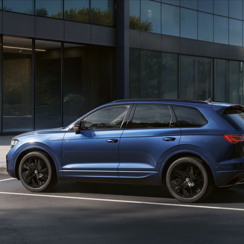 Ein blauer VW Touareg Final Edition steht vor einem Bürogebäude mit Glasfassade - Ansicht seitlich.
