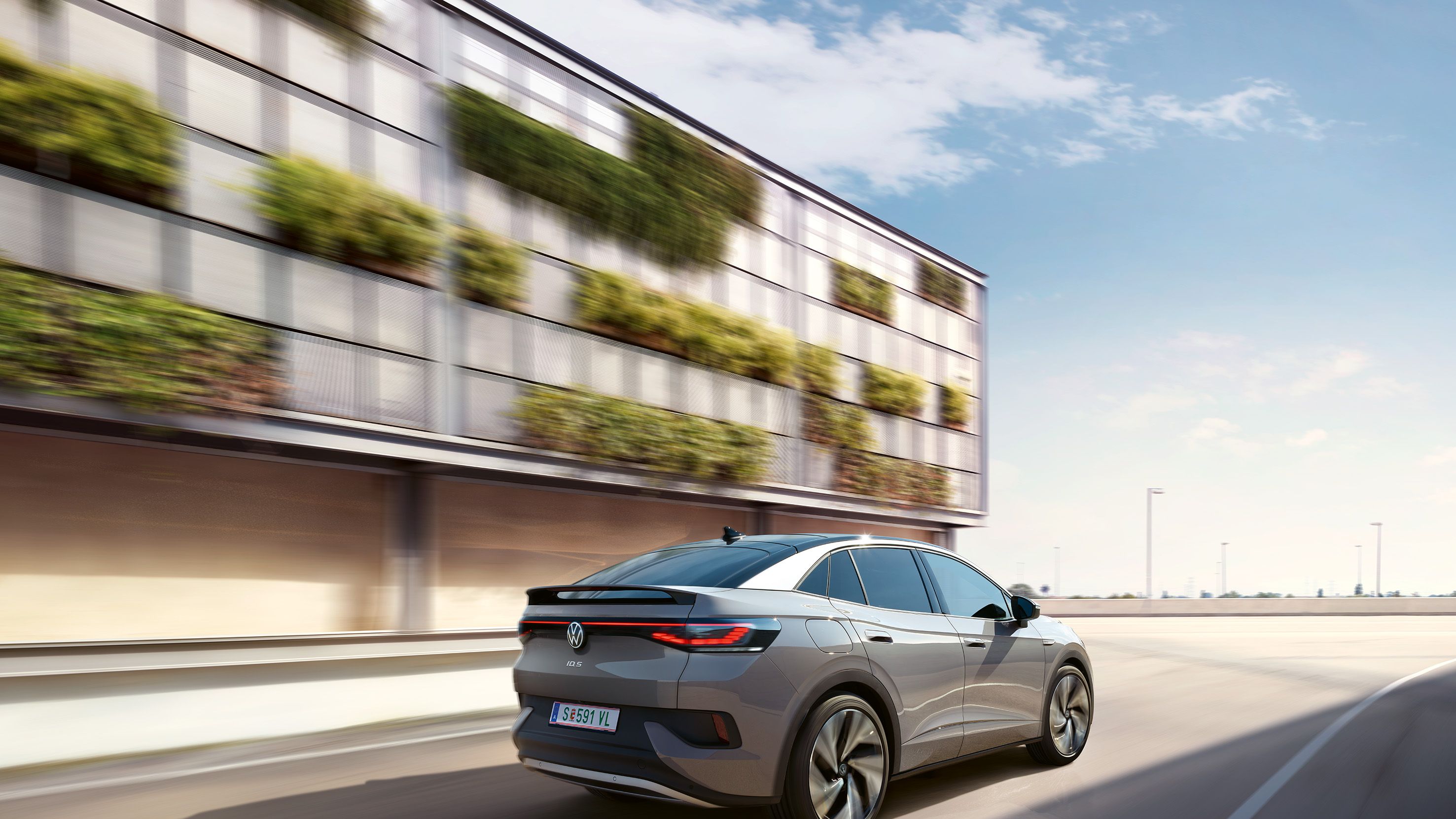 Fahraufnahme eines grauen VW ID.5 von schräg hinten, links ein helles, teilweise grün bewachsenes Gebäude