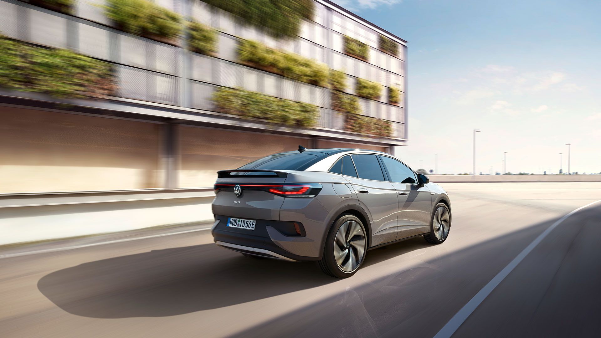 Fahraufnahme eines grauen VW ID.5 von schräg hinten, links ein helles, teilweise grün bewachsenes Gebäude