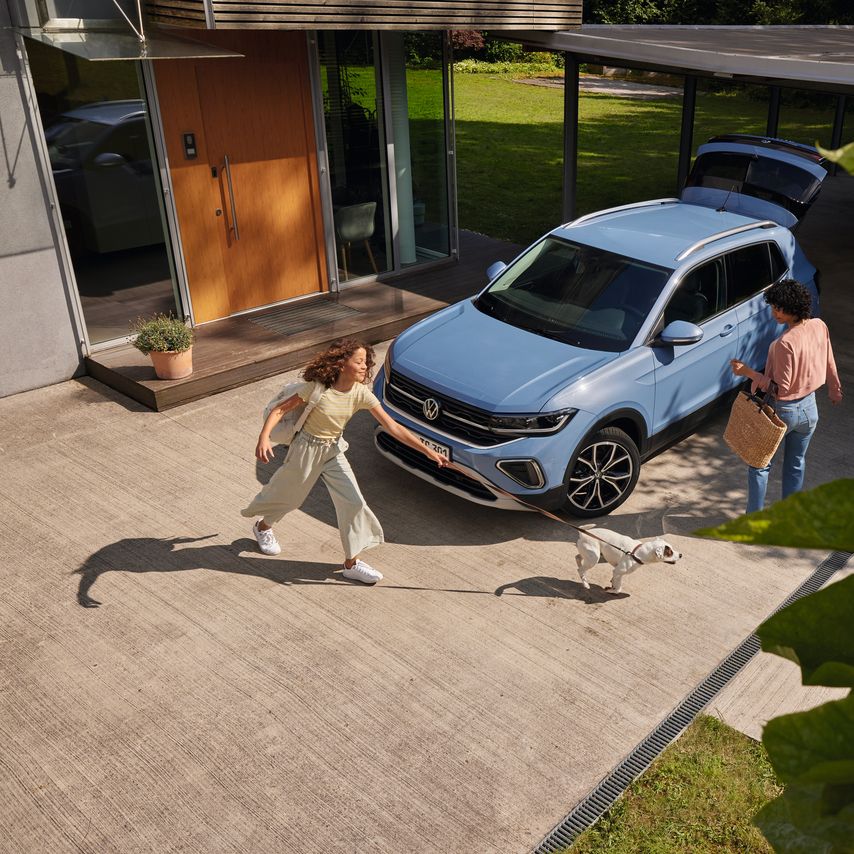 Eine Frau mit Korb und ein Mädchen mit Hund vor einem blauen VW T-Cross mit geöffnetem Kofferraum in einer Einfahrt.