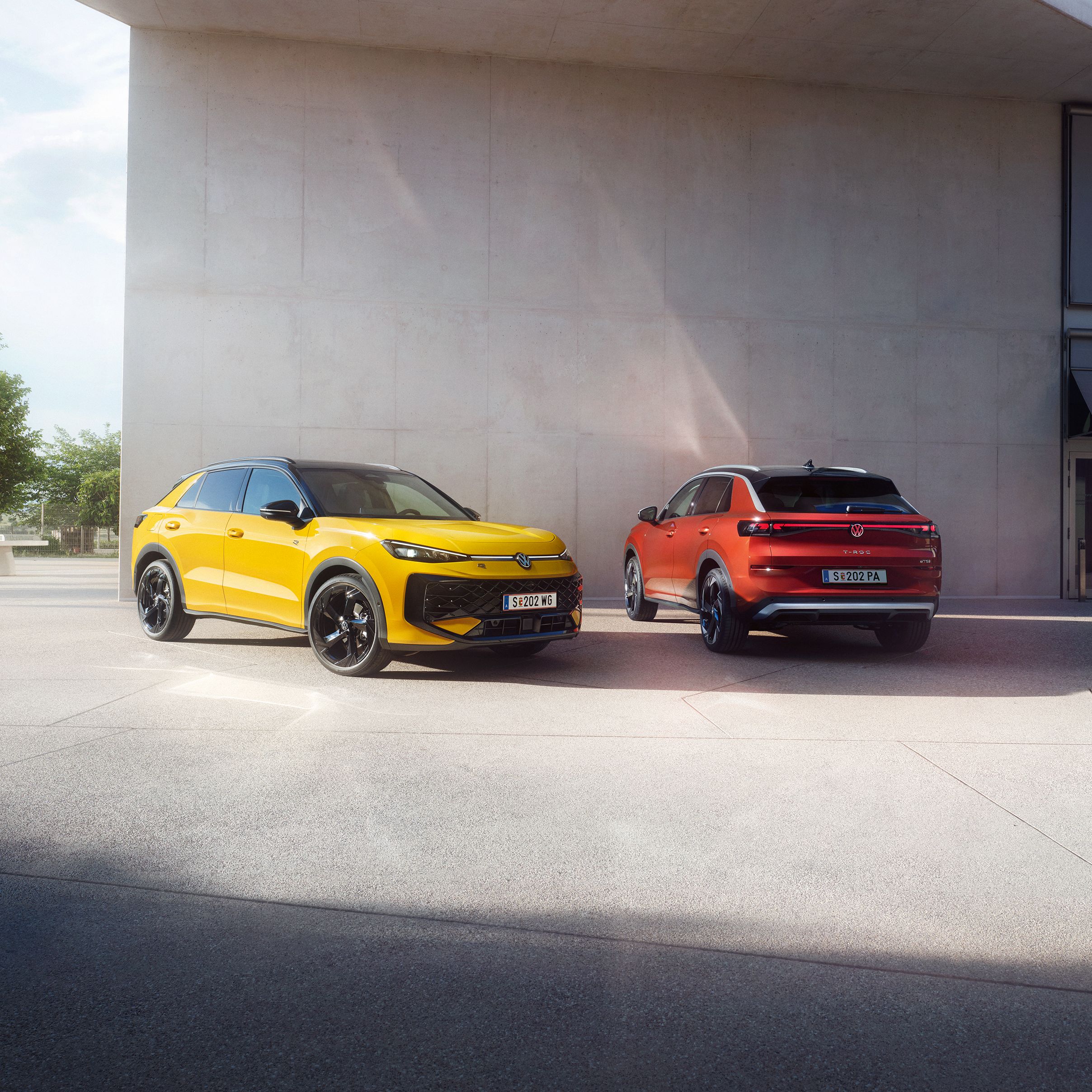 Ein gelber VW T-Roc R-Line und ein roter VW T-Roc Style stehen nebeneinander vor einem modernen Gebäude.