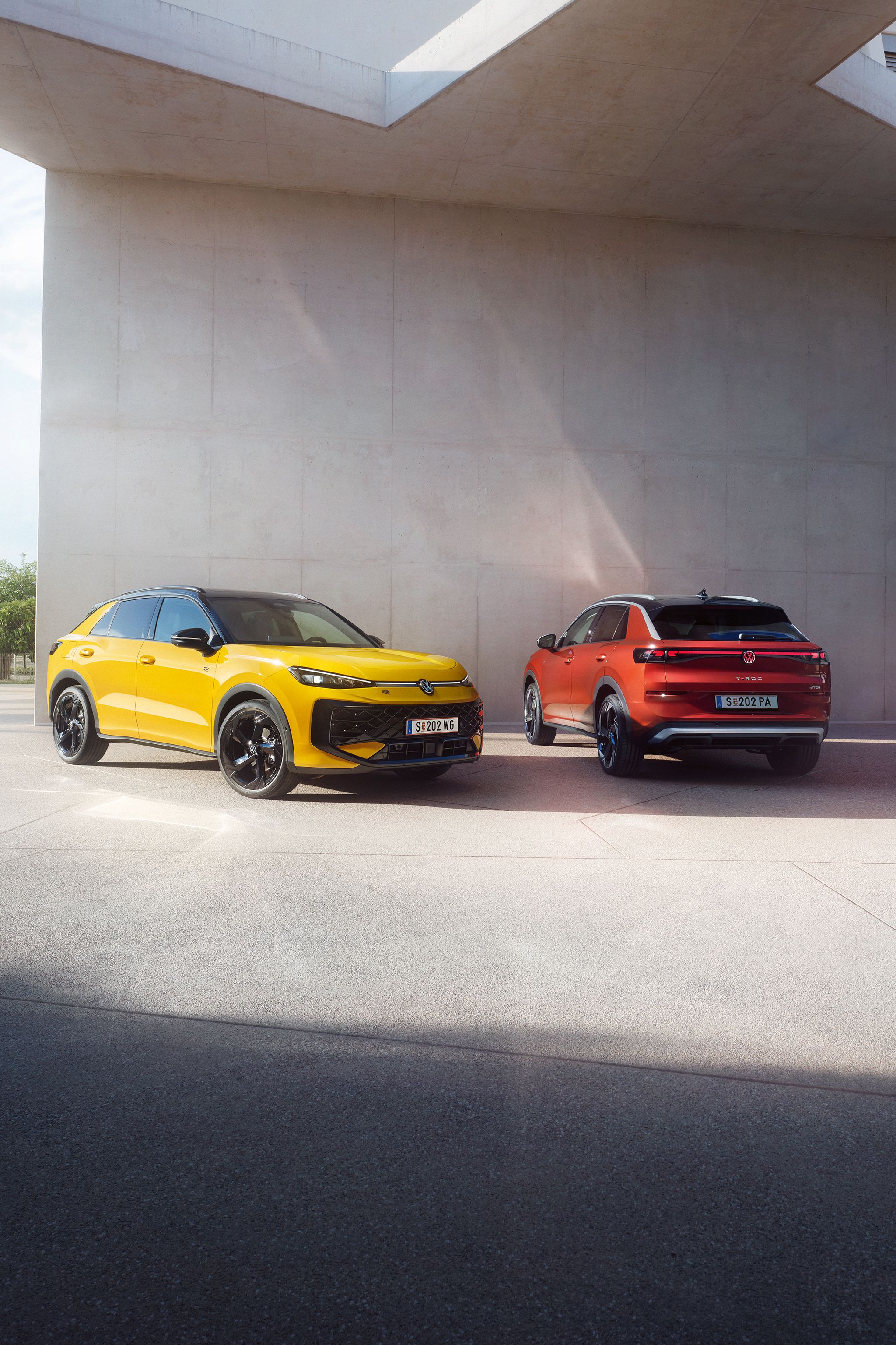 Ein gelber VW T-Roc R-Line und ein roter VW T-Roc Style stehen nebeneinander vor einem modernen Gebäude.