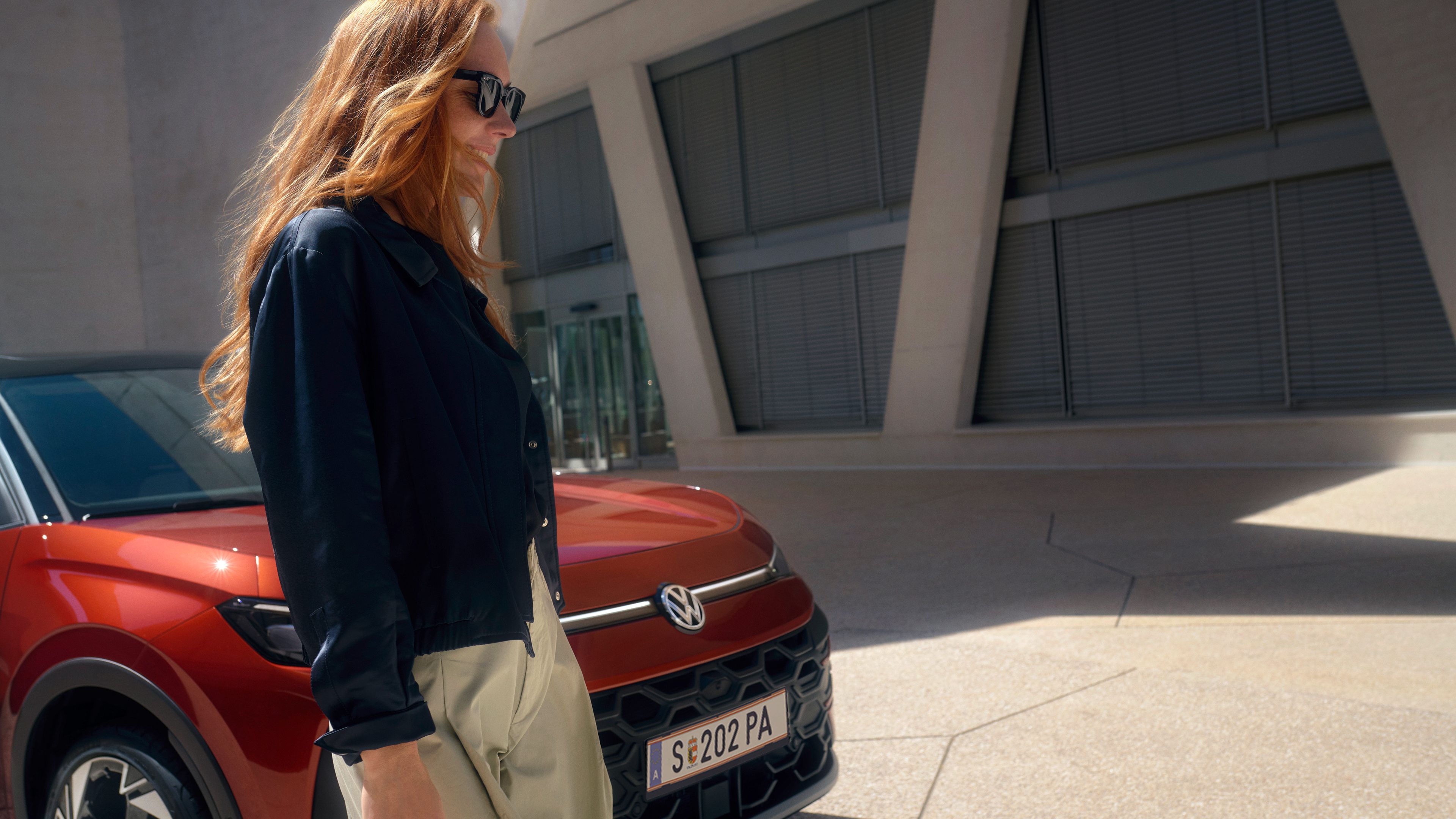 Eine Frau mit Sonnenbrille steht neben einem roten VW TRoc Style vor einem Gebäude.