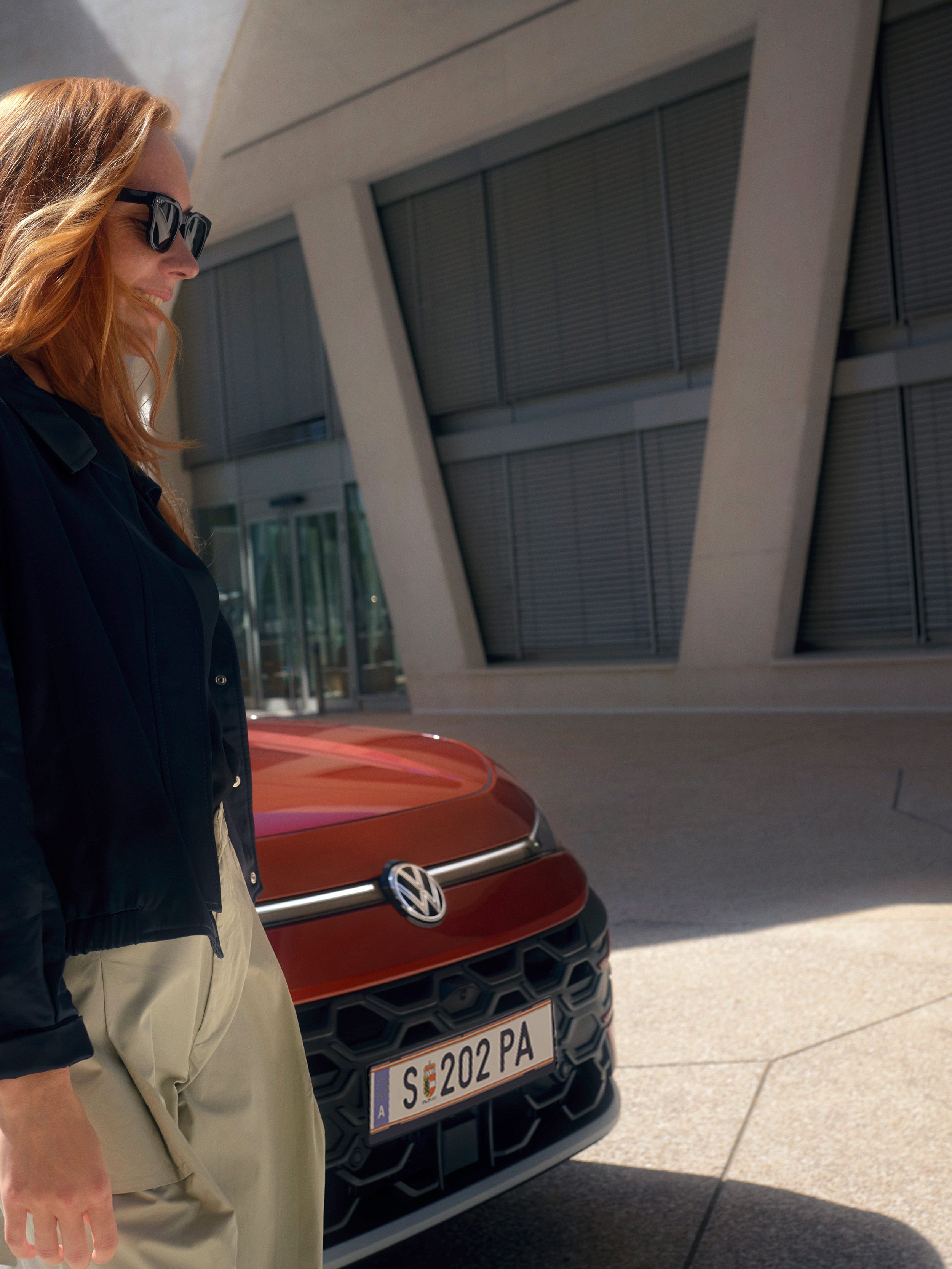 Eine Frau mit Sonnenbrille steht neben einem roten VW TRoc Style vor einem Gebäude.