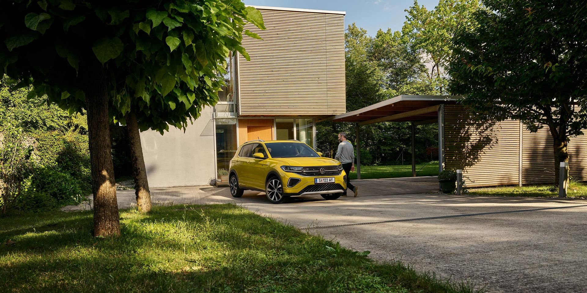 Ein Mann macht sich bereit, in einen gelben VW T-Cross einzusteigen, der in Frontansicht auf einer Garteneinfahrt eines Hauses geparkt ist.