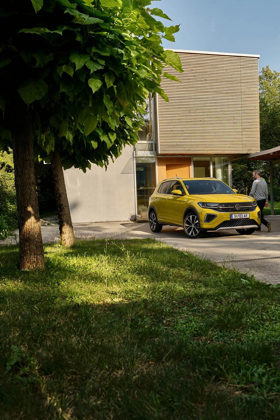 Ein Mann macht sich bereit, in einen gelben VW T-Cross einzusteigen, der in Frontansicht auf einer Garteneinfahrt eines Hauses geparkt ist.