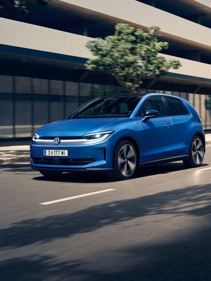 Ein blauer VW ID. Polo Style fährt durch eine Stadt mit grünen Bäumen am Straßenrand.