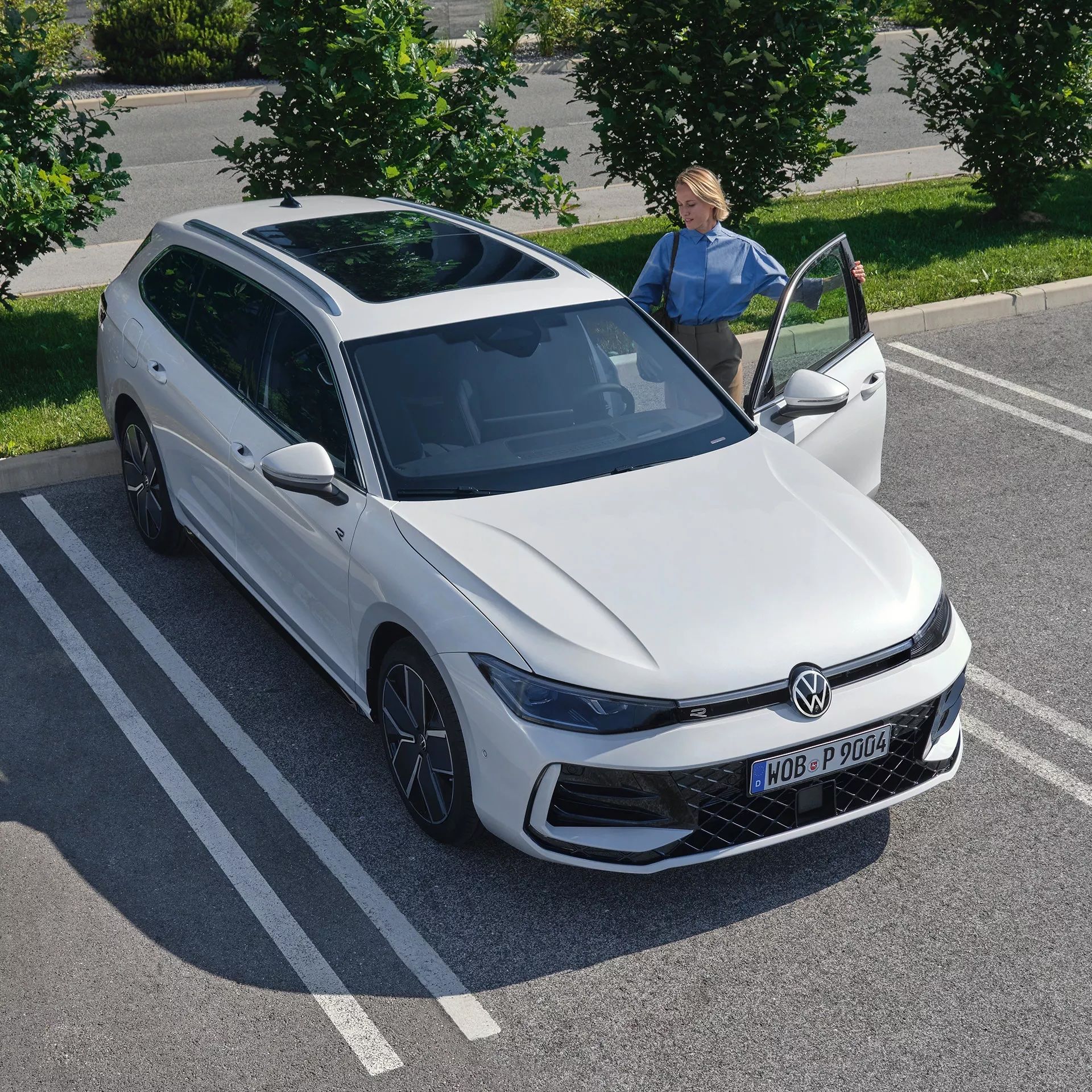 Topshot eines neuen weißen VW Passat auf einem Parkplatz, eine Frau steigt auf der Fahrerseite ein.