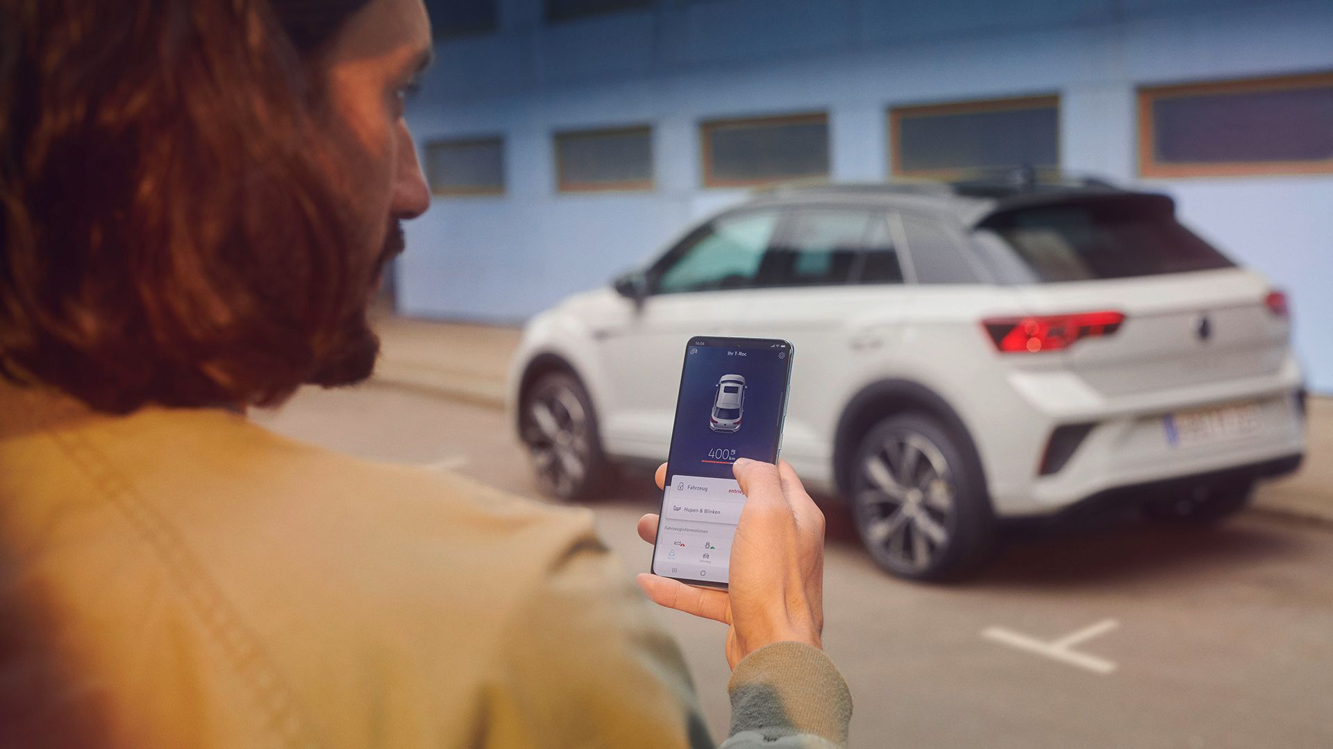 VW T-Roc R-Line in weiß vor einem Gebäude, Seite sichtbar, Mann steht mit Smartphone im Vordergrund