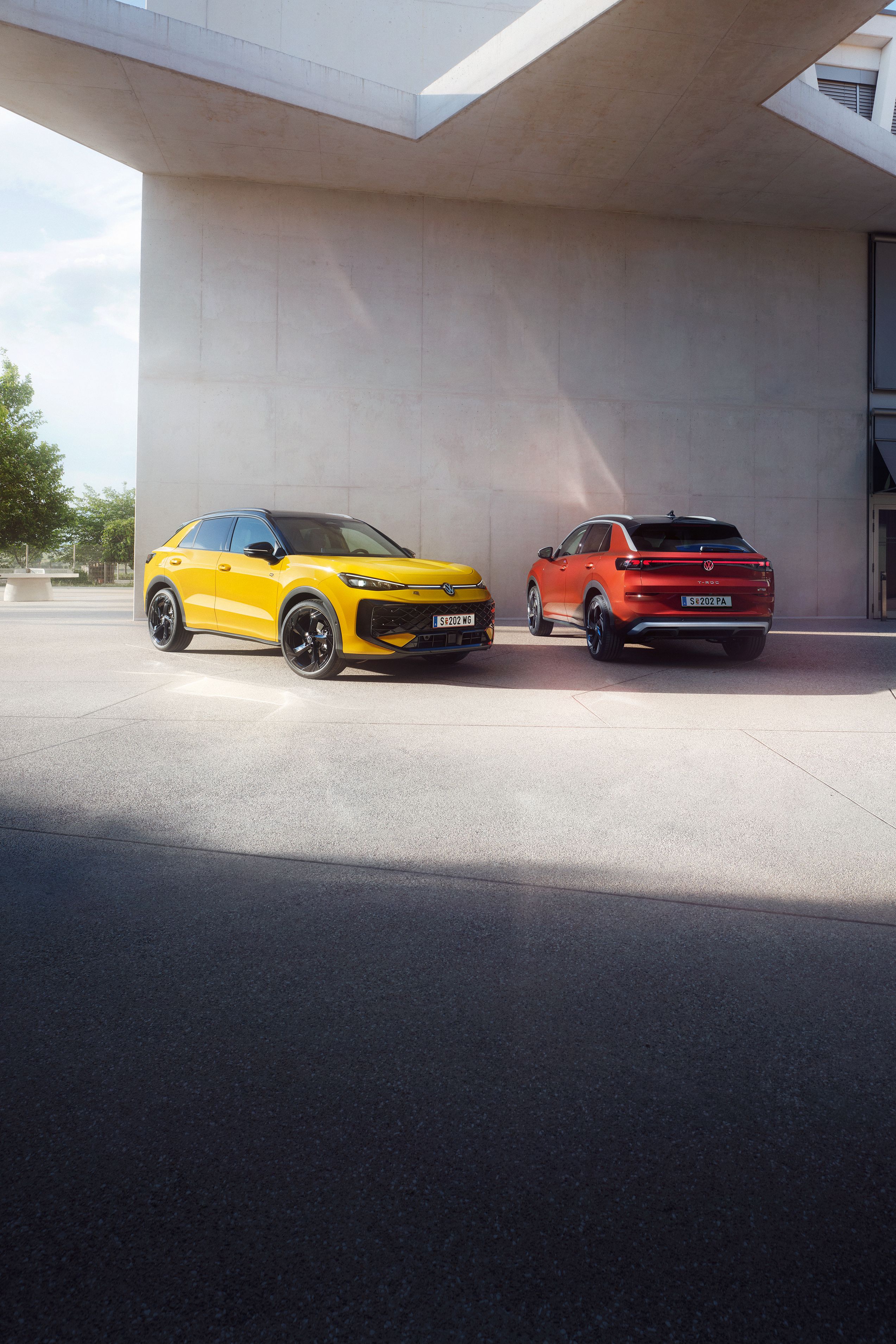 Ein gelber VW T-Roc R-Line und ein roter VW T-Roc Style stehen nebeneinander vor einem modernen Gebäude.