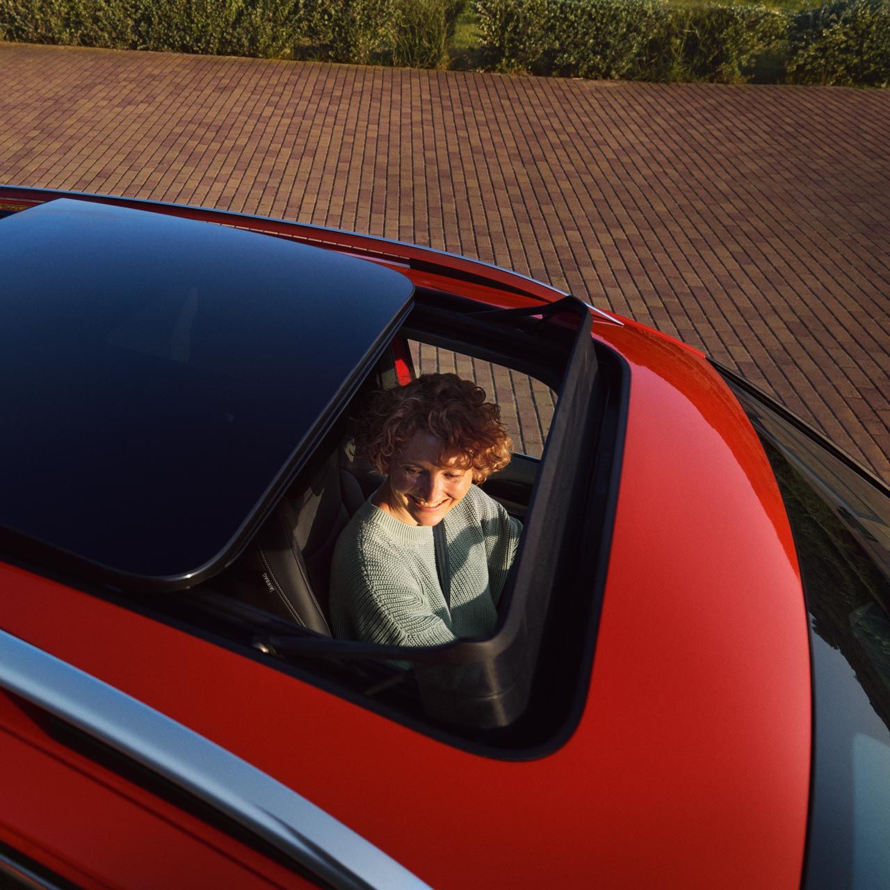 Topshot durch das halb geöffnete optionale Panorama-Ausstell-/ Schiebedach eines roten VW Tiguan. Auf dem Fahrersitz ist eine junge Frau zu sehen.