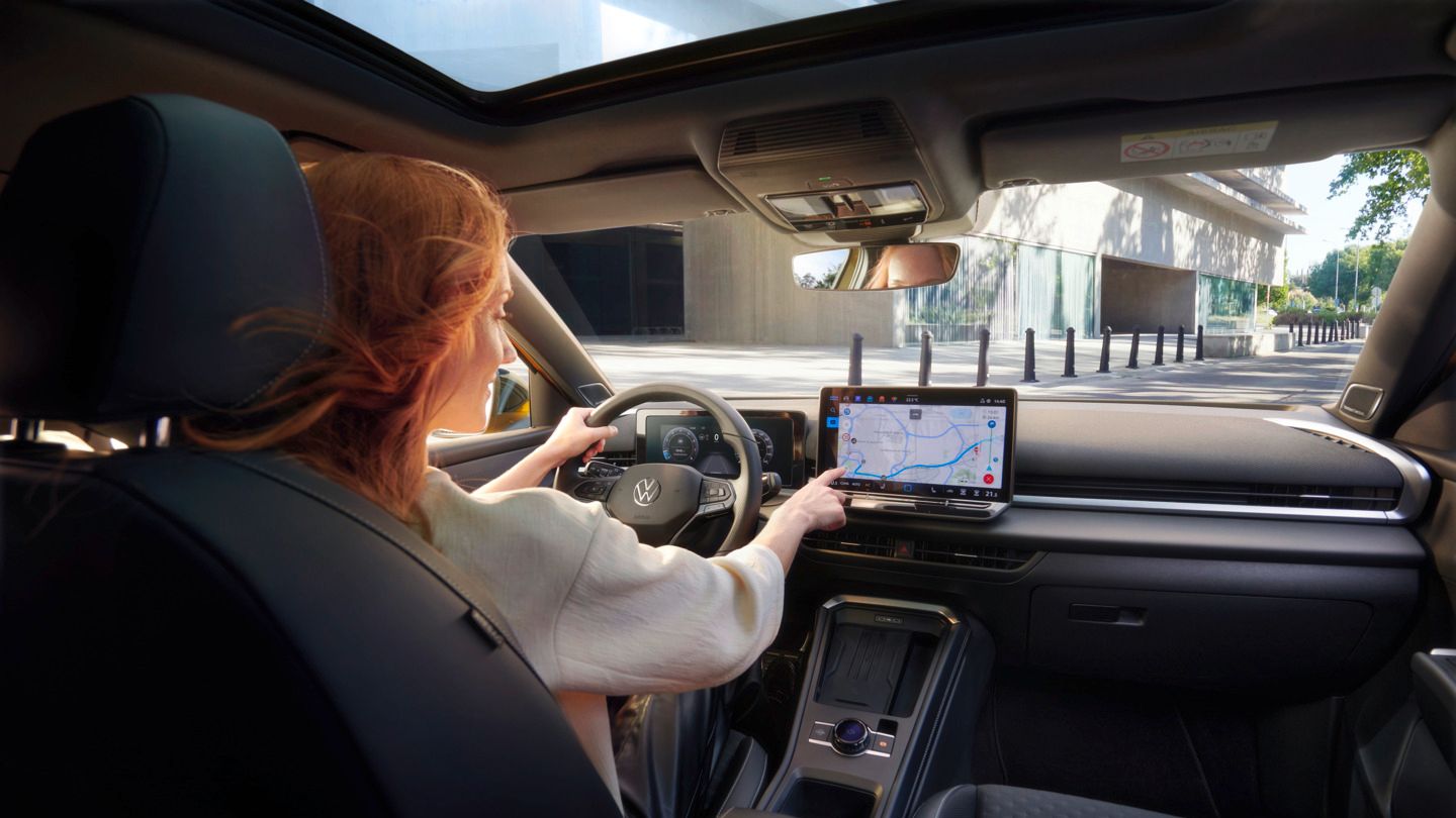 VW T-Roc Eine rothaarige Frau sitzt am Fahrersitz eines VW T-Roc Autos. Man sieht die vordere Hälfte des schwarzen Interieurs. Mit der einen Hand bedient sie die Navigation im Infotainmentsystem per Touchscreen, die andere Hand hat sie auf dem Lenkrad. Das Auto scheint durch eine Stadt zu fahren.