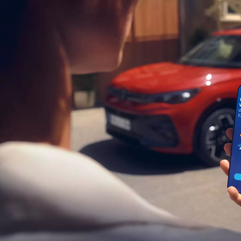 Blick auf ein Handydisplay mit Fahrzeugdaten des Tiguan, im Hintergrund ein seitlich geparkter VW Tiguan in rot
