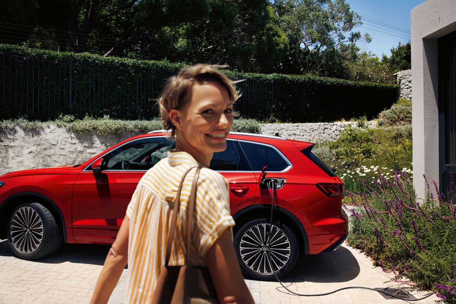 Im Vordergrund eine lächelnde Frau, im Hintergrund lädt ein VW Touareg Elegance eHybrid an einer Ladesäule.
