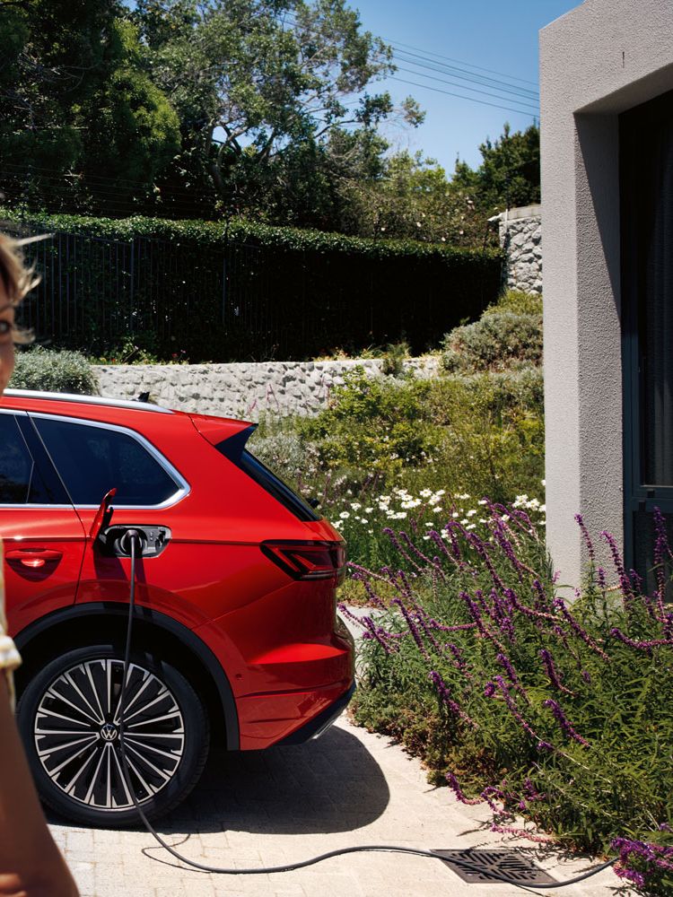 Im Vordergrund eine lächelnde Frau, im Hintergrund lädt ein VW Touareg Elegance eHybrid an einer Ladesäule.