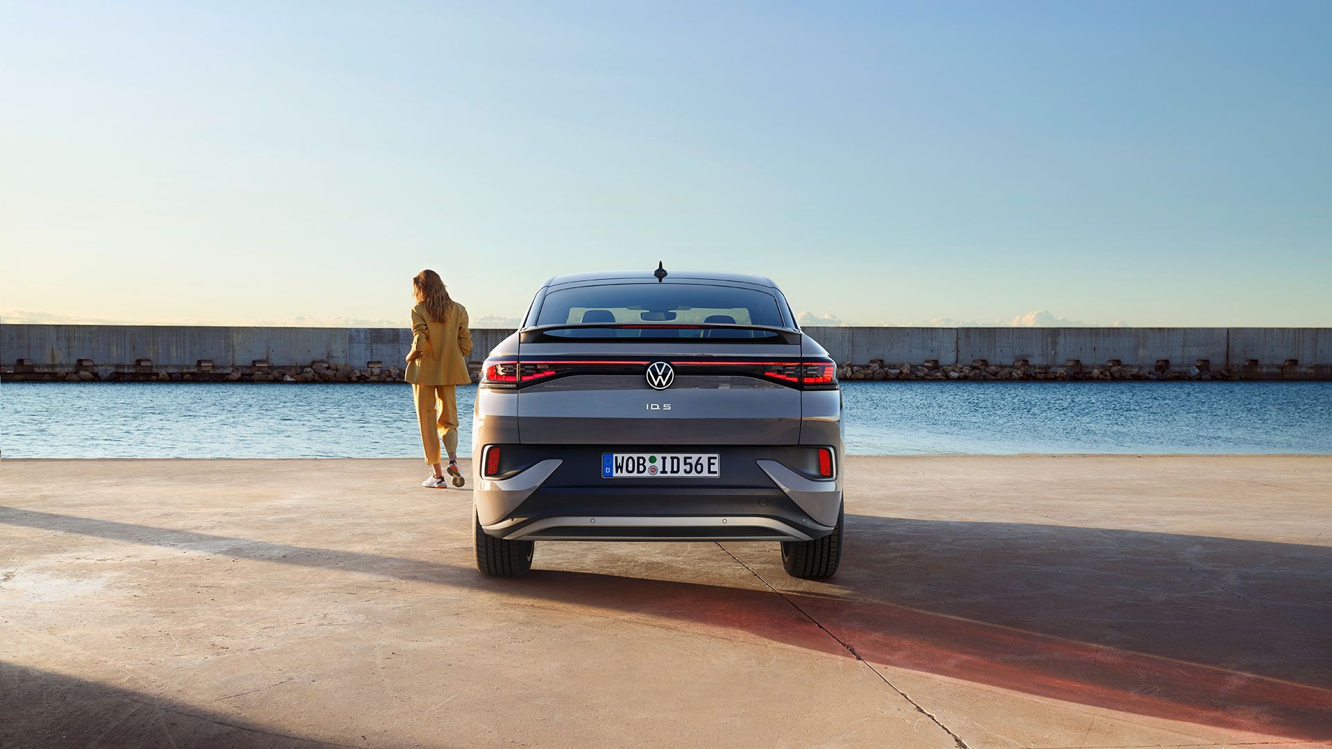 Heckansicht eines VW ID.5 an einem Hafenkai, neben dem E-Auto steht eine Frau