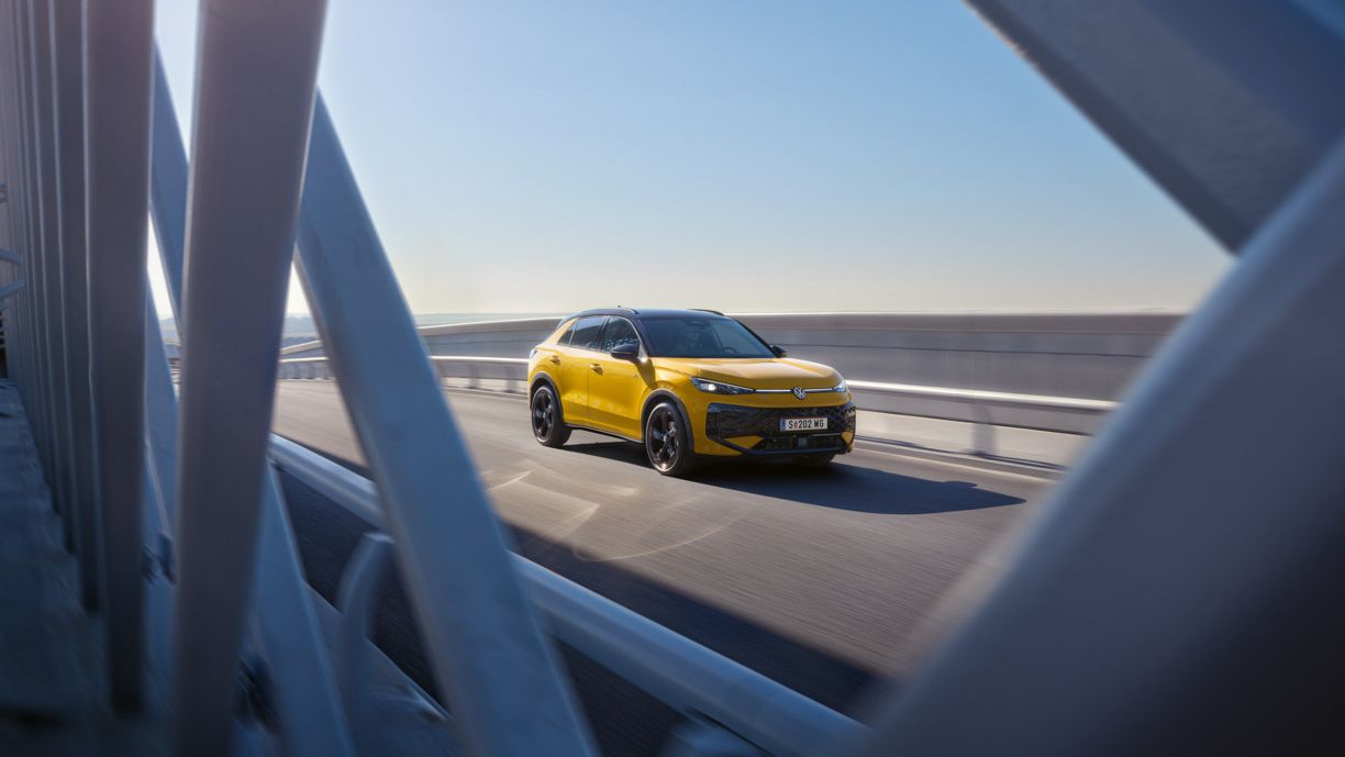 Ein gelber T-Roc fährt über eine moderne Brücke bei klarem Himmel. Die Aufnahme zeigt das Fahrzeug aus der Ferne, teilweise durch die Brückenstrukturen hindurch. Es ist ein sonniger Tag.