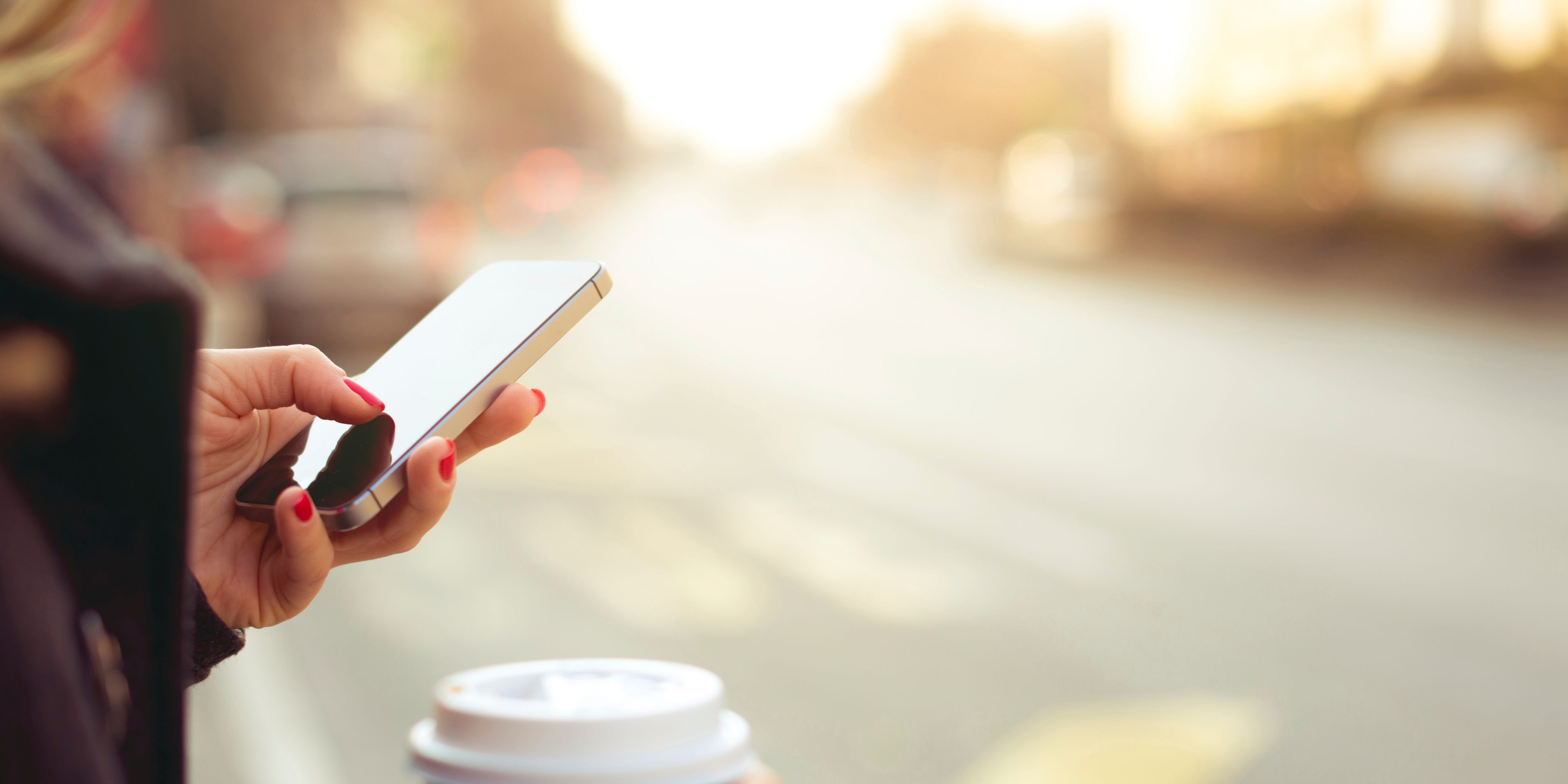 Eine Frau hält in der einen Hand einen Kaffeebecher, mit der anderen Hand bedient sie ihr Smartphone