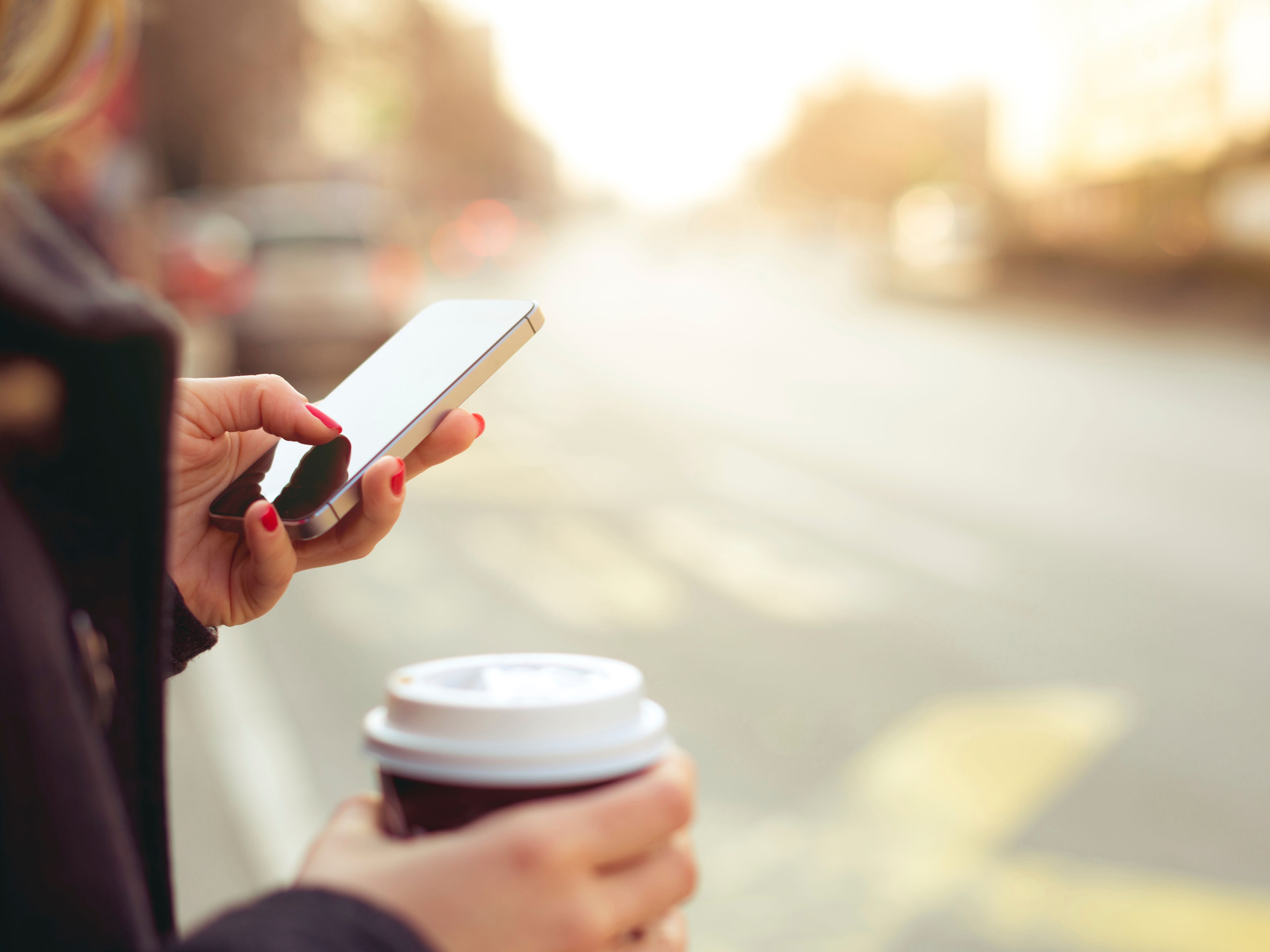 Eine Frau hält in der einen Hand einen Kaffeebecher, mit der anderen Hand bedient sie ihr Smartphone