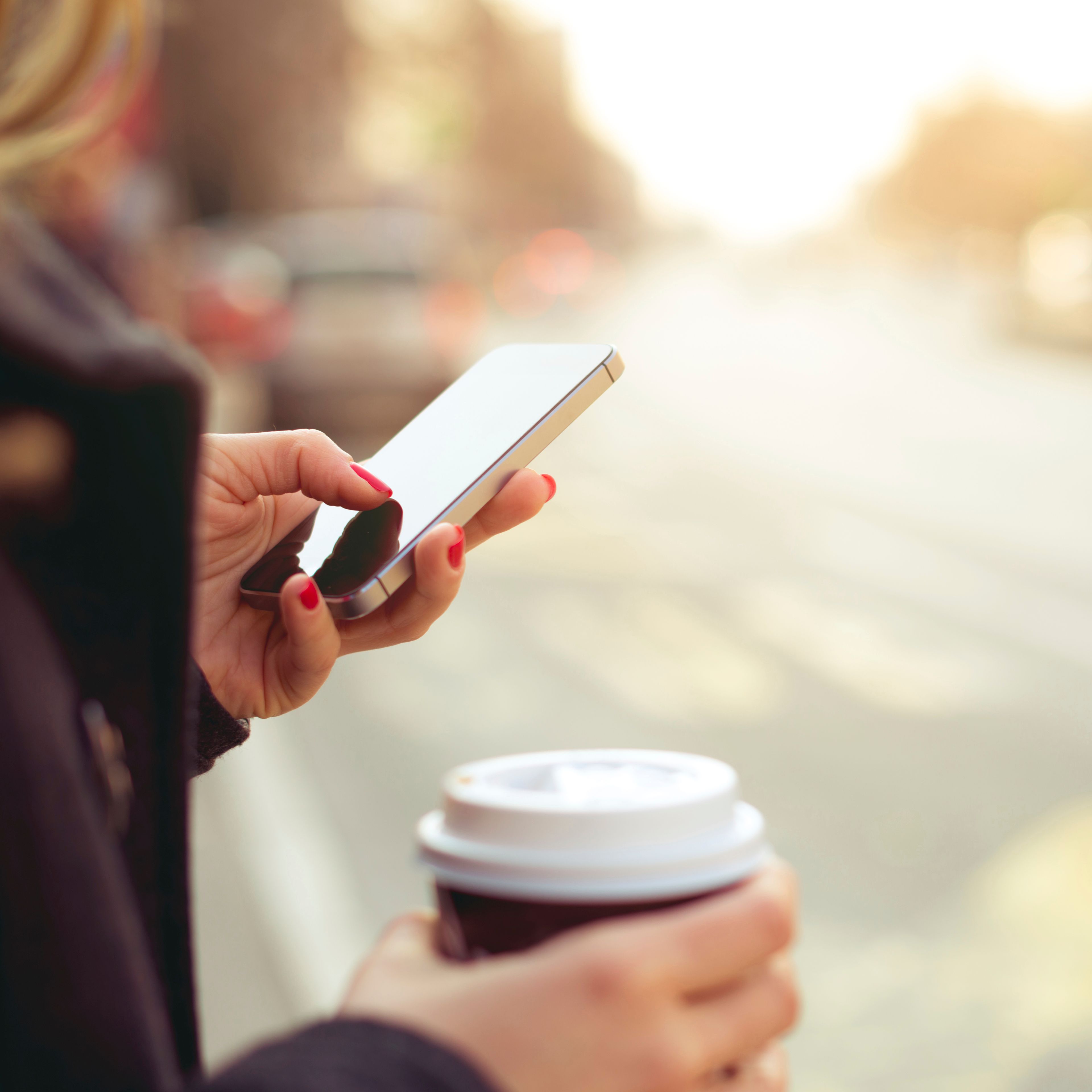 Eine Frau hält in der einen Hand einen Kaffeebecher, mit der anderen Hand bedient sie ihr Smartphone