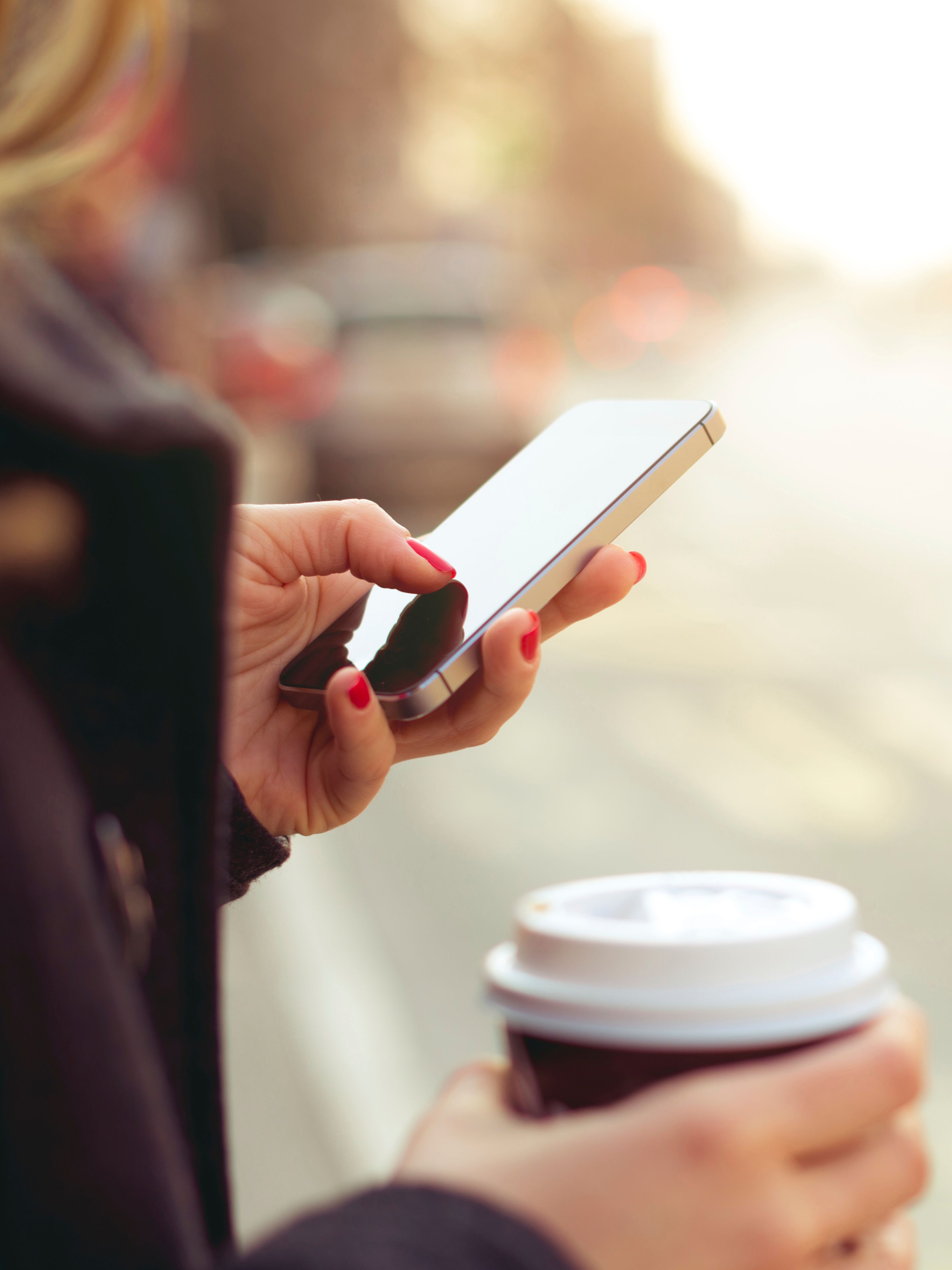 Eine Frau hält in der einen Hand einen Kaffeebecher, mit der anderen Hand bedient sie ihr Smartphone