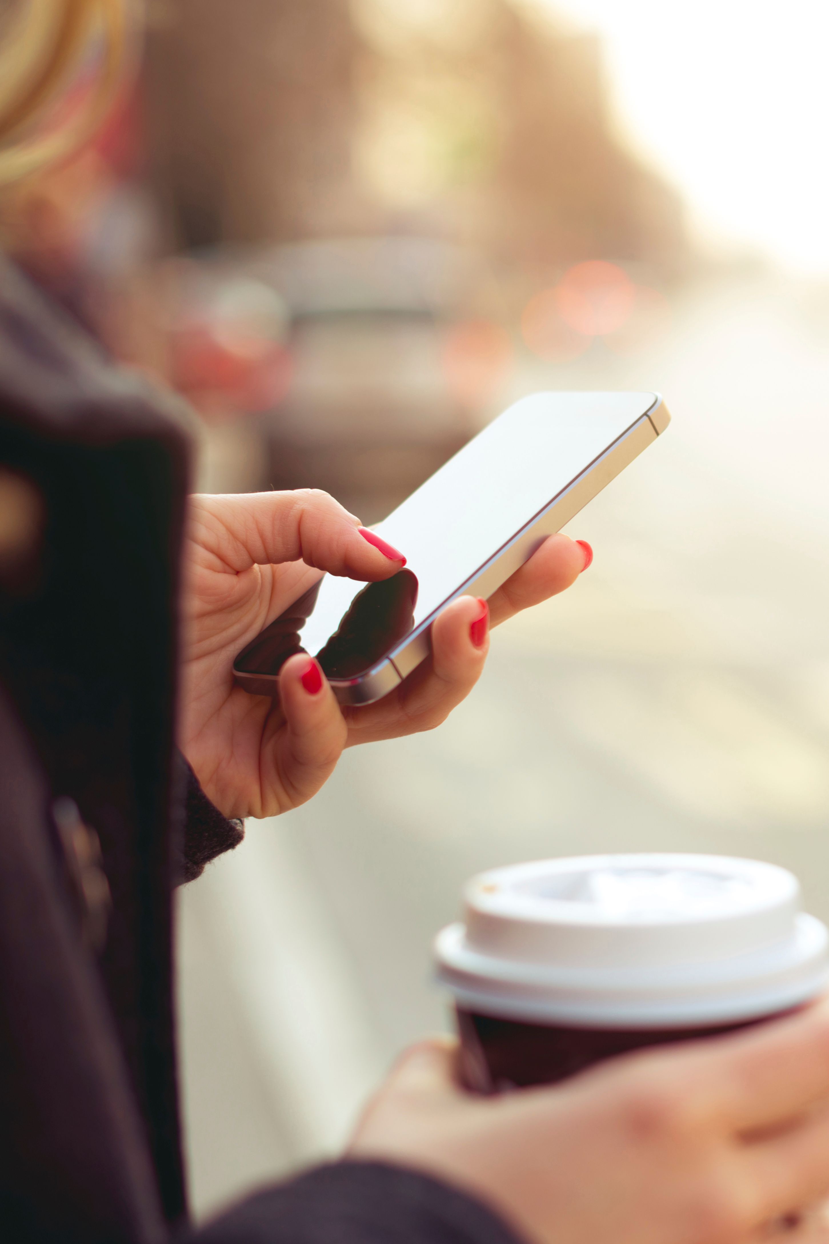 Eine Frau hält in der einen Hand einen Kaffeebecher, mit der anderen Hand bedient sie ihr Smartphone