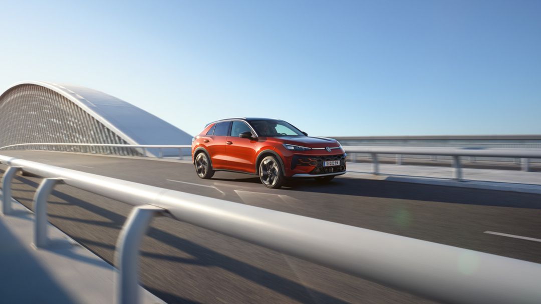 Ein rotfarbener T-Roc fährt über eine moderne Brücke bei klarem Himmel. Die Brücke hat ein großes, gebogenes Element auf der linken Seite. Der Straßenbelag ist sauber und gut erhalten.