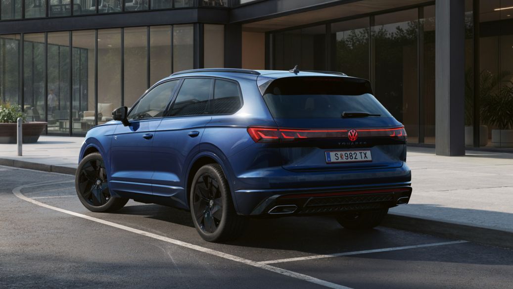 Ein blauer VW Touareg FINAL EDITION  steht vor einem Bürogebäude mit Glasfassade - Ansicht von schräg hinten.