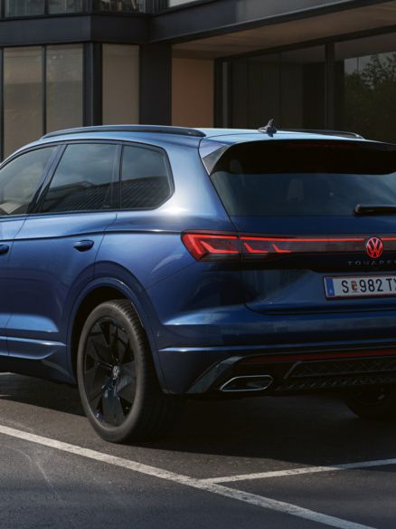 Ein blauer VW Touareg FINAL EDITION  steht vor einem Bürogebäude mit Glasfassade - Ansicht von schräg hinten.