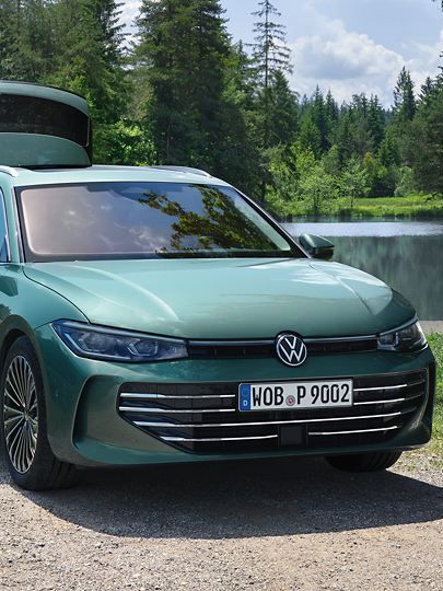 VW Passat in blaugrün parkt an einem Badesee mit geöffnetem Kofferraum. Ein Mann verteilt Freitzeitspielzeug an zwei Kinder.