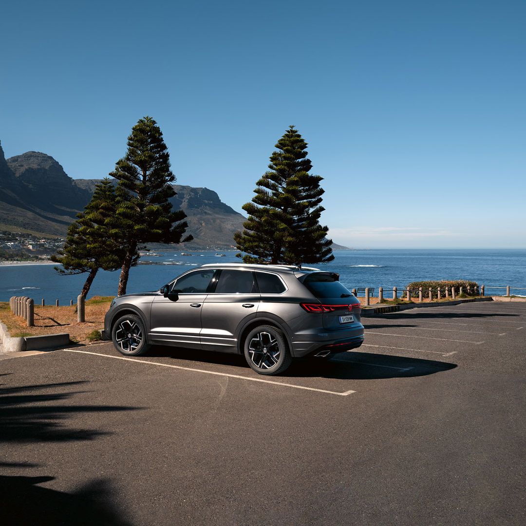 Ein VW Touareg Elegance parkt in der Sonne, Ansicht von der Seite, dahinter sind das Meer und die Küste zu sehen.