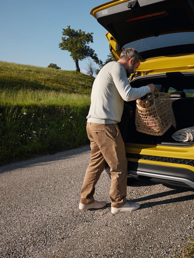 Ein Mann stellt einen großen Picknickkorb in den Kofferraum eines gelben VW T-Cross, der auf einer Straße in hügeliger Landschaft steht.