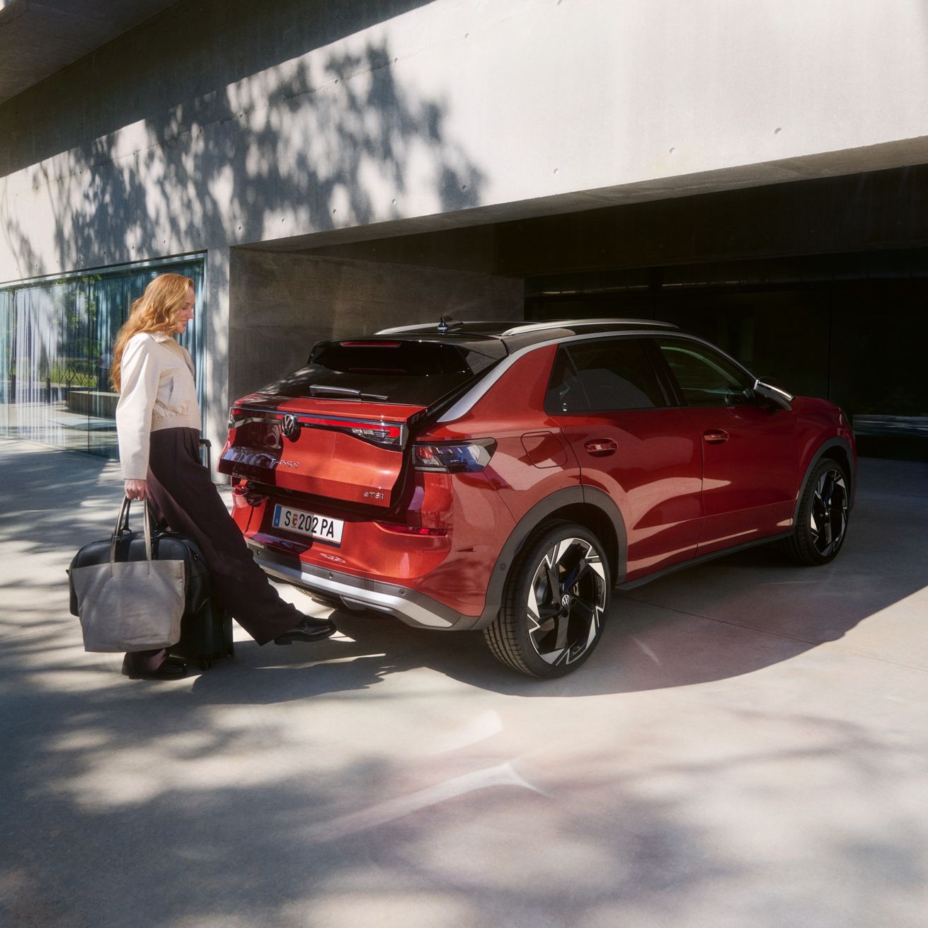 Eine Person mit einer großen Tasche steht hinter einem roten T-Roc SUV, der vor einem modernen Gebäude geparkt ist. Die Frau scheint dabei zu sein, den Kofferraum des Fahrzeugs aufzuschließen.