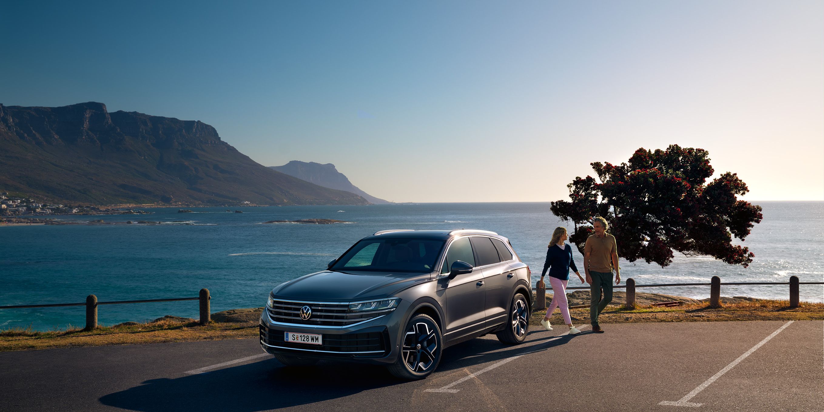Ein VW Touareg Elegance parkt in der Sonne, Ansicht von schräg vorne, dahinter sind das Meer und die Küste zu sehen.