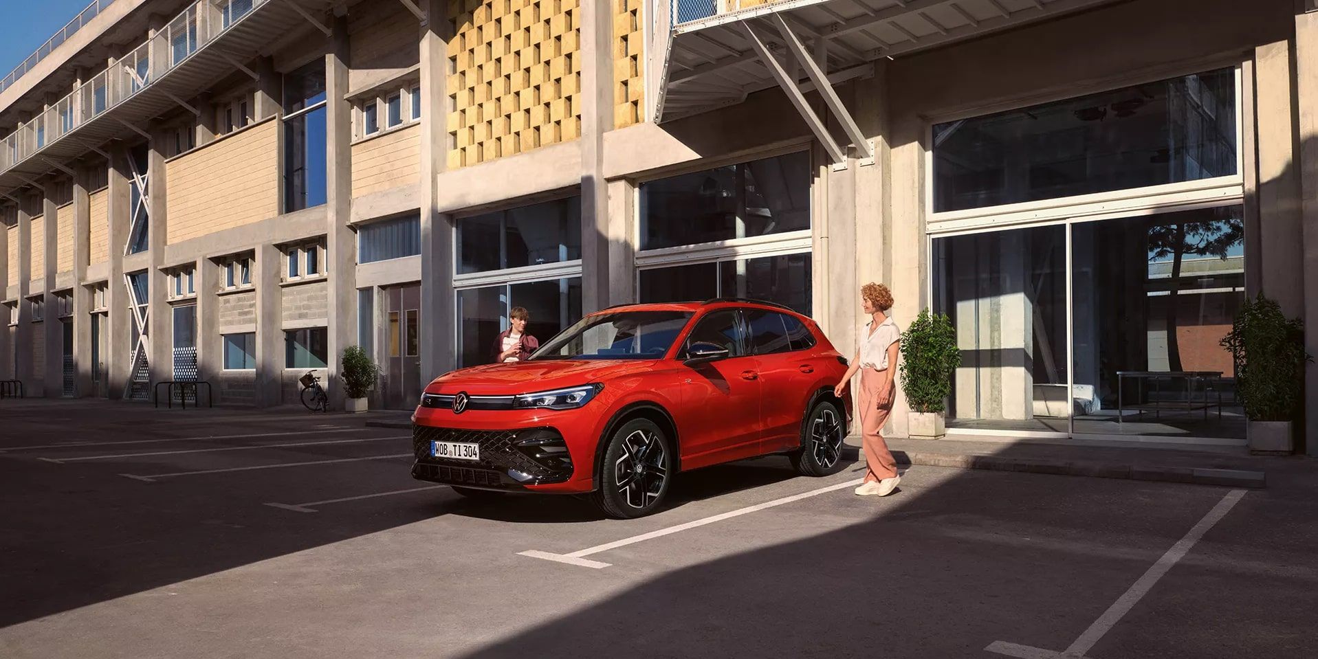 Roter VW Tiguan in Seitenansicht steht vor einem Gebäude, eine junge Frau geht auf der Fahrerseite vorbei.