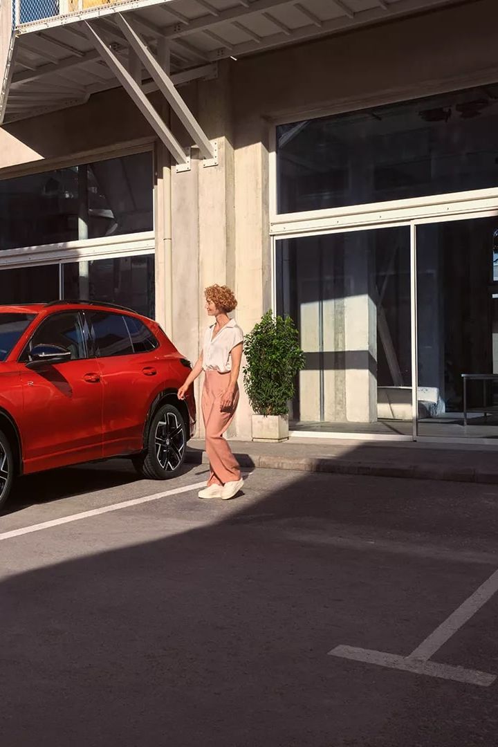 Roter VW Tiguan in Seitenansicht steht vor einem Gebäude, eine junge Frau geht auf der Fahrerseite vorbei.