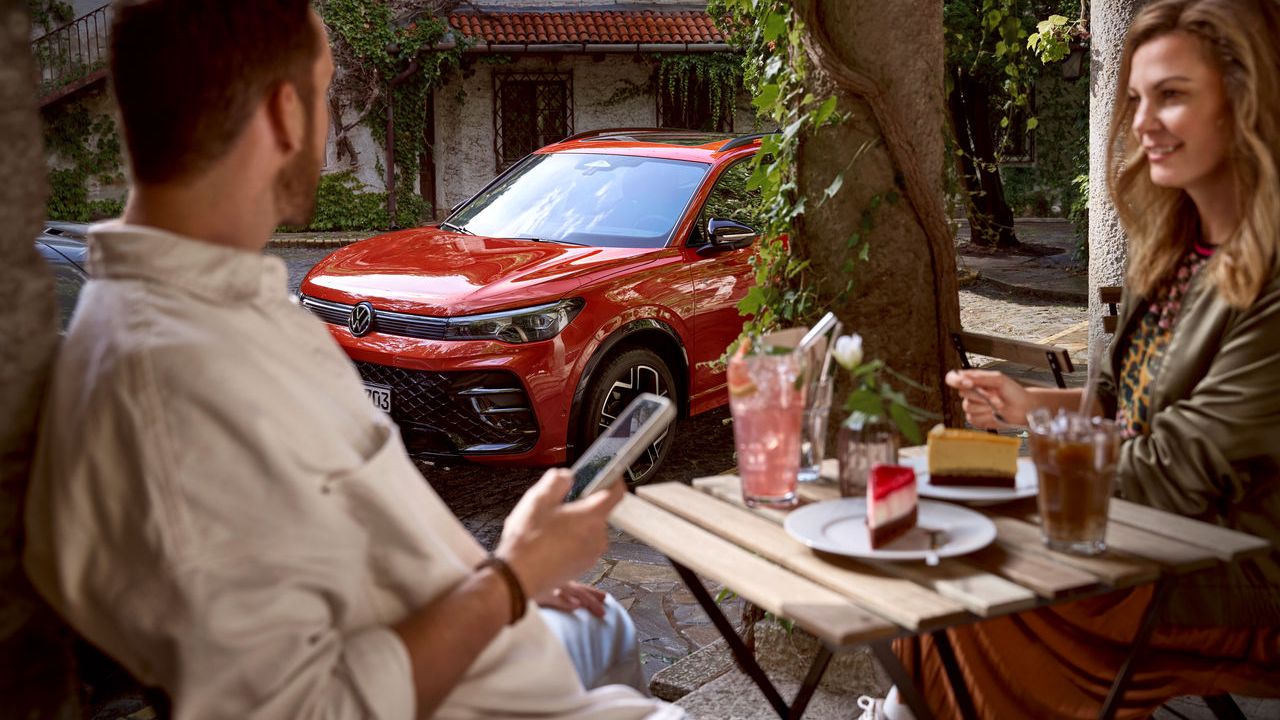 Mann und Frau sitzen in einem Kaffee und der Mann hält sein Handy in der Hand und schaut auf sein Tiguan