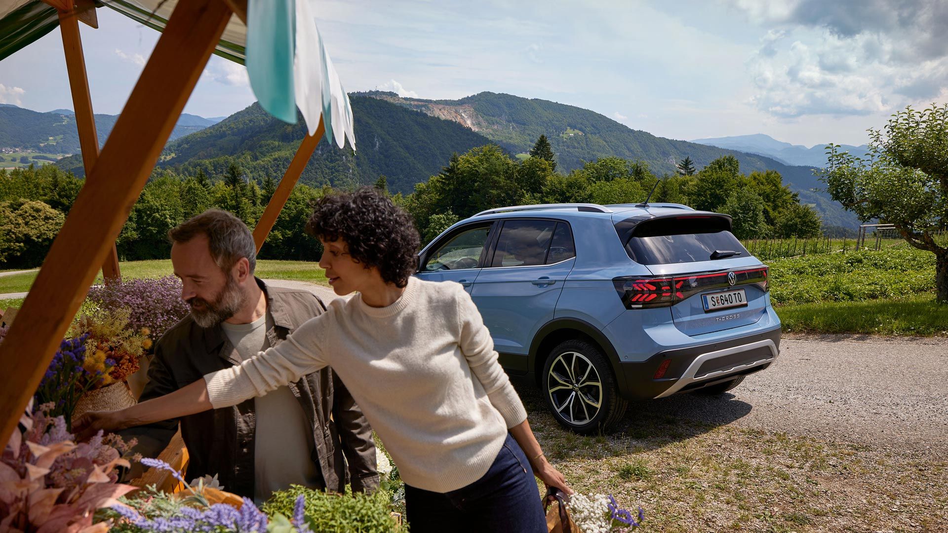 Mann und Frau stehen bei einem Blumenstand, hinter ihnen ein blauer VW T-Cross vor bergiger Landschaft.