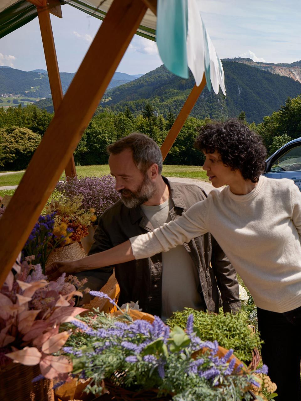 Mann und Frau stehen bei einem Blumenstand, hinter ihnen ein blauer VW T-Cross vor bergiger Landschaft.