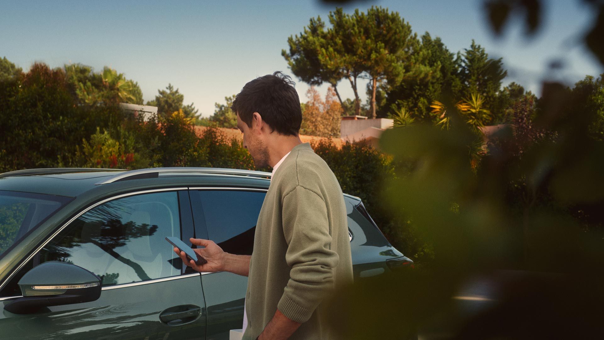 Ein junger Mann auf der Fahrerseite des dunkelgrünen VW Tiguan bedient den optionalen Park Assist Plus über sein Smartphone.