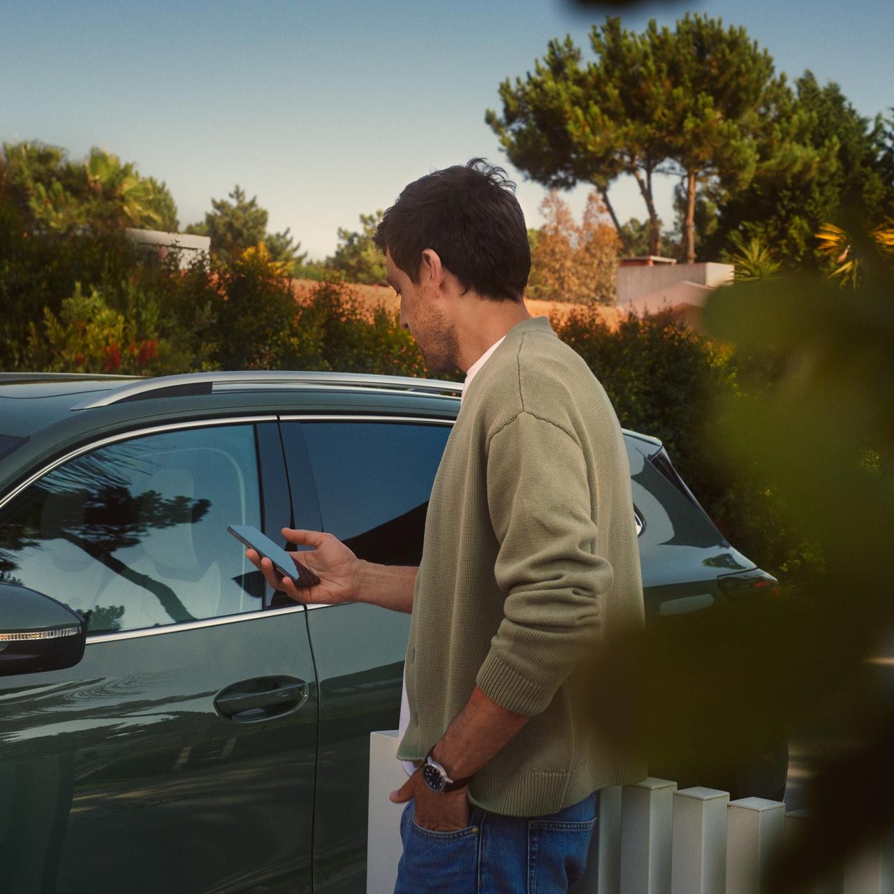 Ein junger Mann auf der Fahrerseite des dunkelgrünen VW Tiguan bedient den optionalen Park Assist Plus über sein Smartphone.