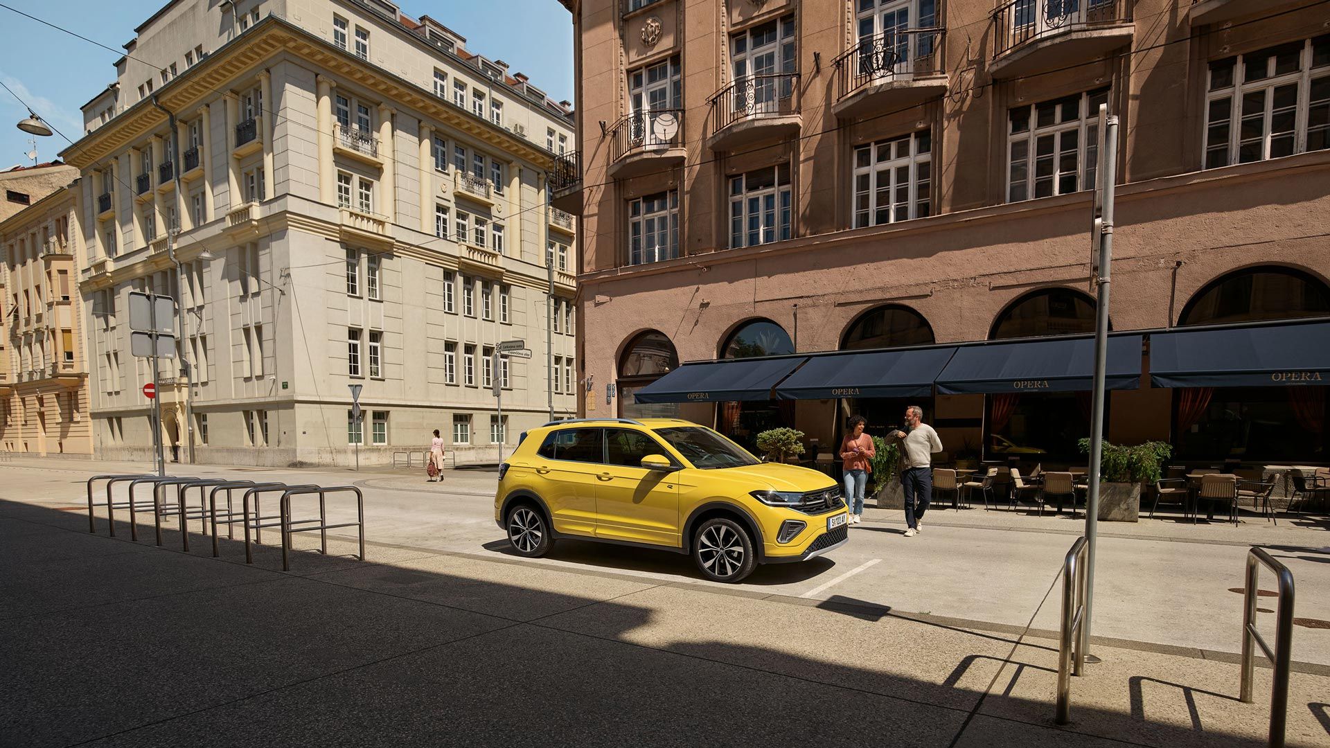 VW T-Cross in gelb parkt seitlich vor einem urbanen Straßencafé, ein Päarchen geht auf den Wagen zu.