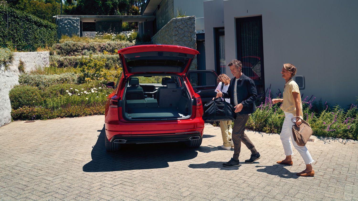 Ein Mann und eine Frau gehen auf einen VW Touareg Elegance eHybrid mit geöffnetem Kofferraum zu.