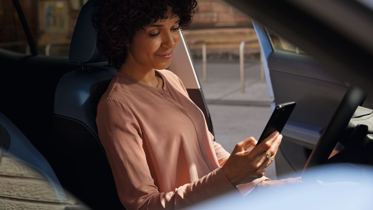 Eine lächelnde Frau schaut auf dem Fahrersitz eines parkenden VW T-Cross aufs Handy.