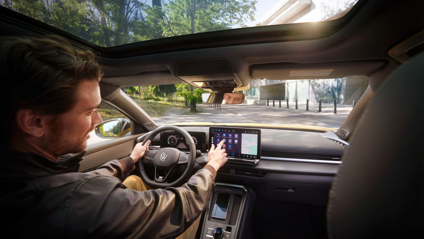 VW T-Roc Ein Mann mit braunen Haaren und schwarzer Jacke sitzt am Fahrersitz im Interieur eines VW T-Rocs und bedient das Infotainmentsystem und mit der anderen Hand das Lenkrad. Er befindet sich auf einer Straße, im Hintergrund sieht man rechts ein modern wirkendes Gebäude, links ein bewaldetes Stück.