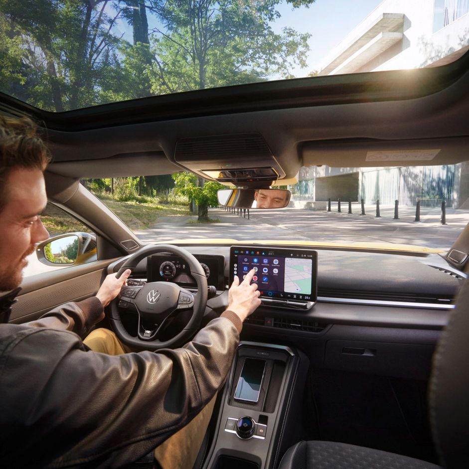 Ein Mann mit braunen Haaren und schwarzer Jacke sitzt am Fahrersitz im Interieur eines VW T-Rocs und bedient das Infotainmentsystem und mit der anderen Hand das Lenkrad. Er befindet sich auf einer Straße, im Hintergrund sieht man rechts ein modern wirkendes Gebäude, links ein bewaldetes Stück.