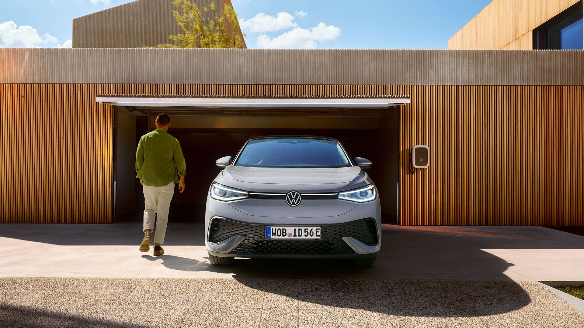 Grauer ID.5 steht in Einfahrt einer Garage. Daneben der ID.Charger am Haus. Mann geht in Garage.