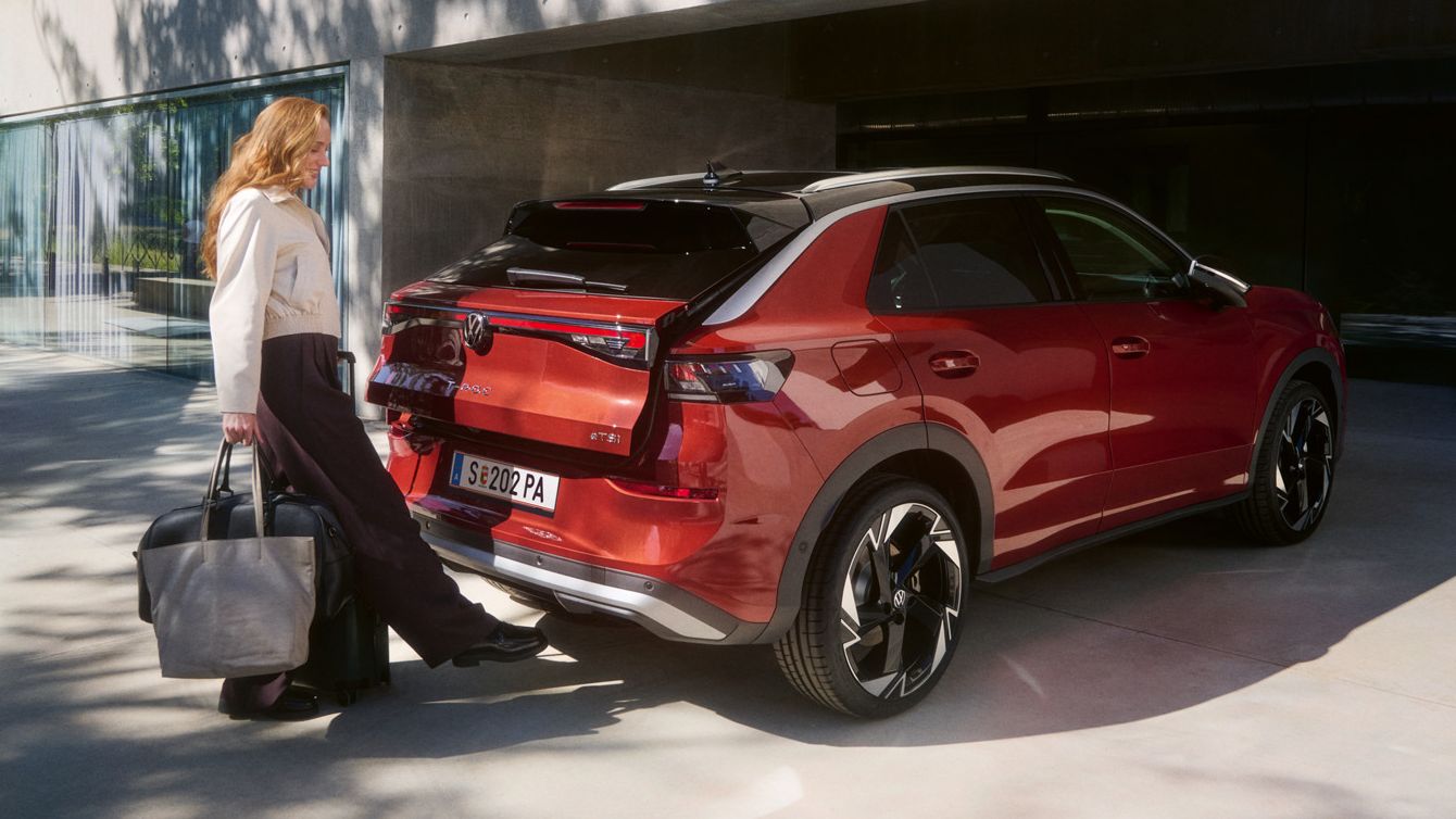 Eine Person mit einer großen Tasche steht hinter einem roten T-Roc SUV, der vor einem modernen Gebäude geparkt ist. Die Frau scheint dabei zu sein, den Kofferraum des Fahrzeugs aufzuschließen.